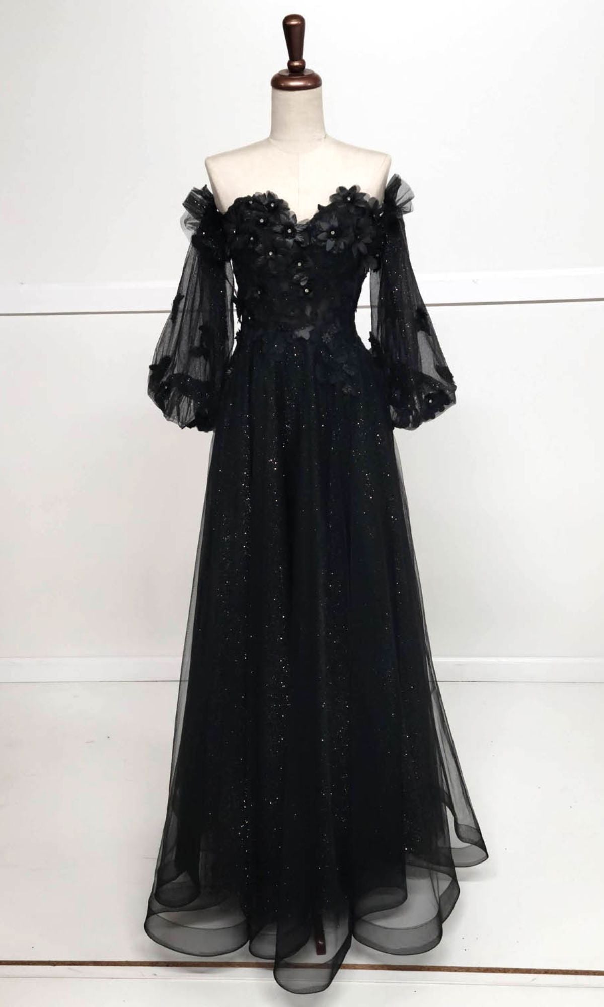 The Chicas CHF3096 is a black, off-the-shoulder prom ball gown with sheer puff sleeves, a floral applique bodice, and a floor-length glittery tulle skirt displayed on a dress form.